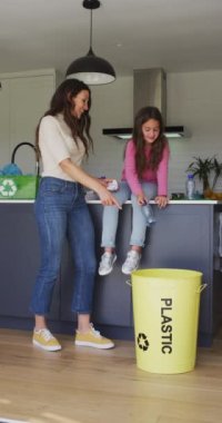 Kafkasyalı bir anne ve kızın mutfakta atık ayıklarken çekilmiş dikey bir fotoğrafı, plastik bir su şişesini bir kaba atan bir kız. Aile ve geri dönüşüm kavramı.