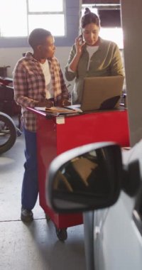 Laptop ve akıllı telefon kullanan iki farklı kadın araba tamircisinin dikey videosu. Araba tamirhanesinde çalışmak ve küçük kadınsı iş konsepti yürütmek..