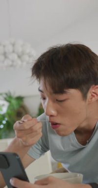 Asyalı bir gencin oturma odasında kahvaltı edişinin dikey videosu. Evde yalnız zaman geçirmek..