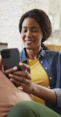 Kanepede oturan ve akıllı telefon kullanan mutlu Afro-Amerikan kadının dikey videosu. Rahatlayın, rahatlayın ve evde teknolojiyle vakit geçirin..