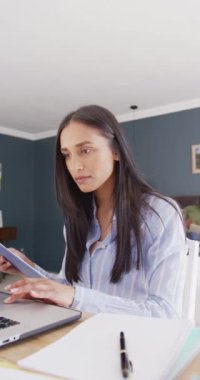 Yatak odasında dizüstü bilgisayar kullanan ve notlar okuyan melez bir kadının dikey videosu. İletişim, evden çalışma, ev hayatı ve kapsayıcılık kavramı.