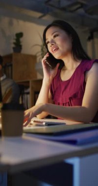 Asyalı iş kadınının akıllı telefon kullandığı, geç saatlere kadar çalıştığı dikey bir video. İş ve ofiste geceleri teknoloji konseptiyle çalışmak.