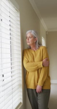 Düşünceli, son sınıf öğrencisi bir kadının dikey videosu. Evinde durup pencereden dışarı bakıyor. Emeklilik, ev hayatı ve yalnızlık kavramı.