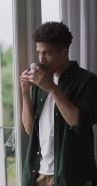 Pencereden dışarı bakıp kahve içen düşünceli melez bir adamın dikey videosu. ev hayatı, evde yalnız zaman geçirmek..