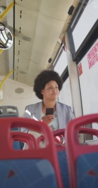 Otobüste akıllı telefon kullanan mutlu ve çift ırklı bir kadının dikey videosu. Şehirde yaşıyor ve ayak işlerini yapıyor..