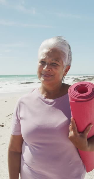 日当たりの良いビーチでヨガマットと幸せな出産の女性の肖像画の垂直ビデオ 健康的で活発な退職ビーチの休日 — ストック動画