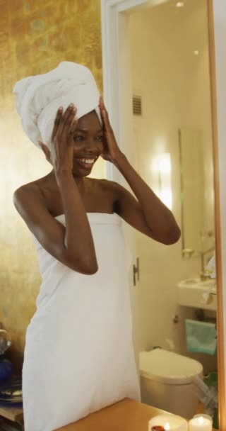 垂直录像中 一个身穿毛巾的非洲裔美国女人微笑着 对着镜子拍拍自己的脸 家庭生活方式 在家里享受温泉休闲时间 — 图库视频影像