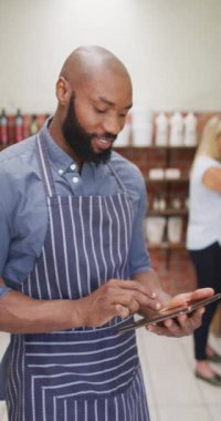 Önlüklü gülümseyen Afro-Amerikan 'ın organik dükkanda tablet kullandığının dikey videosu. Çevre alışverişi, organik tarım ve taze organik sebzeler..