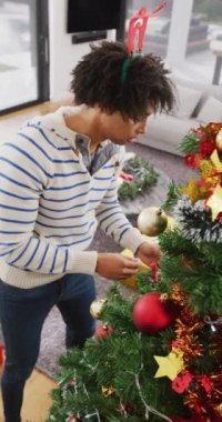 Vertical video of happy biracial man in reindeer antlers decorating christmas tree at home. Christmas, celebration, happiness, tradition and inclusivity concept.