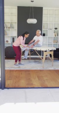 Üst düzey hastayla egzersiz yapan mutlu çift ırklı kadın fizyoterapistin dikey videosu. Yaşlılar sağlık ve huzurevi konsepti.