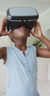 Evde VR kulaklığı kullanan mutlu Afrikalı Amerikalı kızın dikey videosu. çocukluk yaşam tarzı, bedensel bütünlük, teknolojiyle evde keşfin tadını çıkarmak..