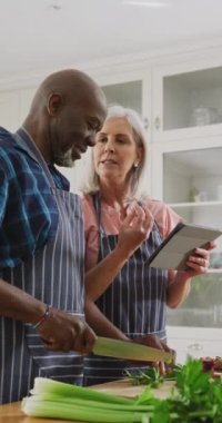 Mutfakta tablet kullanarak yemek hazırlayan mutlu yaşlı çiftin dikey videosu. İletişim, mutluluk, sağlık, emeklilik, ev hayatı ve kapsayıcılık kavramı.