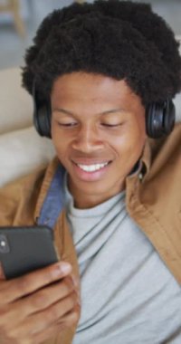 Kulaklık takmış, akıllı telefondan müzik dinleyen mutlu Afro-Amerikan bir adamın dikey videosu. ev hayatı, beden katılımı, teknolojiyle evde kaliteli zaman geçirmek..