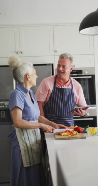 Son sınıf beyaz bir çiftin mutfakta yemek pişirirken, sebze keserken ve tablet kullanırken dikey bir fotoğrafı. Yaşlılar ve ev hayatı kavramı.