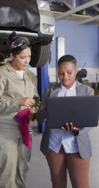 Dizüstü bilgisayar kullanan iki farklı kadın araba tamircisinin dikey videosu. Araba tamirhanesinde çalışmak ve küçük kadınsı iş konsepti yürütmek..