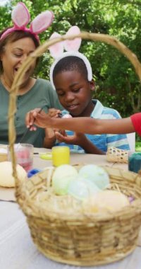 Vertical video of happy african american family painting eggs at easter. family spending time together at home.