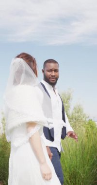 Vertical video of happy diverse bride and groom walking holding hands and talking at outdoor wedding. Marriage, love, happiness and inclusivity concept.