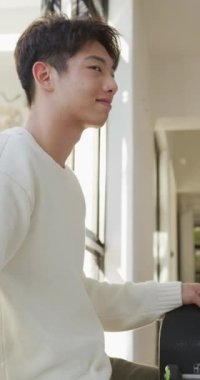 Asyalı bir gencin oturma odasında kaykayla gülümseyen portresinin dikey videosu. Evde yalnız zaman geçirmek..