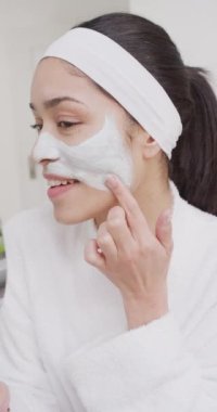 Çift ırklı bir kadının banyoda yüzüne krem sürdüğü dikey bir video. Güzellik, sağlık ve kadın spa konsepti.