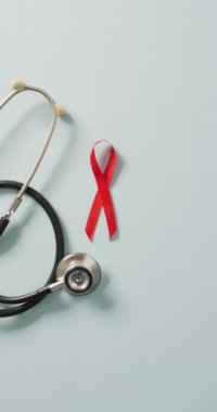 Steteskopun dikey videosu ve gri arka planda kırmızı kurdele. Sağlık, önleme, ilaç, semboller ve kanser farkındalığı kavramı.