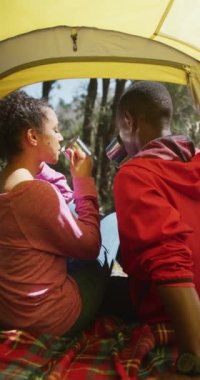 Gülümseyen Afro-Amerikan çiftlerin çadırda oturup kahve içmelerini gösteren dikey bir video. sağlıklı, aktif açık hava yaşam tarzı, romantizm ve boş zaman.