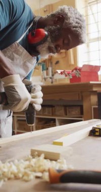 Kıdemli Afro-Amerikan erkek marangozun atölyede sondaj yaptığı dikey video. zanaat, ahşap işçiliği ve küçük bağımsız işletme.