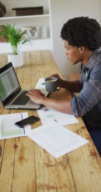 Afro-Amerikan bir adamın kahve içerken dizüstü bilgisayar ve akıllı telefon kullandığını gösteren dikey bir video. evcil yaşam tarzı, bedensel kapsayıcılık, evden teknoloji ile çalışmak.