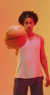 Turuncu arka planda topu olan çift ırklı erkek basketbolcunun dikey videosu. Spor ve rekabet konsepti.