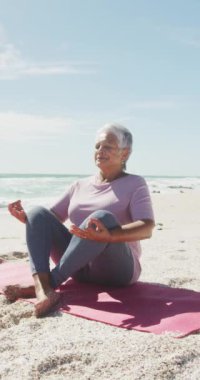 Sunny Beach 'te meditasyon yapan mutlu, melez bir kadının dikey videosu. sağlıklı, aktif emeklilik plaj tatili.