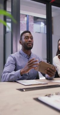 Afro-Amerikan bir adamın elinde tabletle iş yerinde iş arkadaşlarına seslenişini gösteren dikey bir video. Yaratıcı bir işte, iş yerinde bedensel bütünlük.