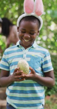 Vertical video of portrait of happy african american boy with egg at easter. spending time at home.