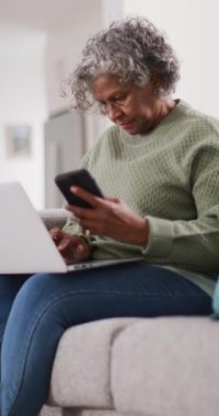 Kanepede dizüstü bilgisayar ve akıllı telefon kullanarak oturan mutlu Afrikalı Amerikalı kadının dikey videosu. Emeklilik ve ev kavramında zaman geçirme.