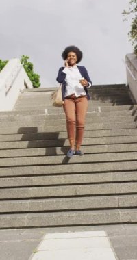 Afro-Amerikan kadının merdivenlerden elinde kahveyle indiği ve akıllı telefondan konuştuğu dikey bir video. Vücut kapsamı, teknoloji gidiyor, şehir dışında ve etrafında.