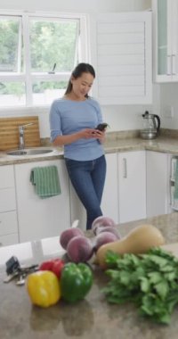 Gülümseyen Asyalı kadının akıllı telefon kullanarak mutfakta durduğu dikey video. ev hayatı, evdeki teknolojinin keyfini çıkarmak..