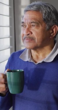 Camdan dışarı bakıp evde kahve içen düşünceli, melez bir adamın dikey videosu. Emeklilik, sağlık, kapsayıcılık ve yaşlılar yaşam tarzı konsepti.