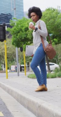 Şehirde yürüyen, meyve suyu içen, dizüstü bilgisayar tutan mutlu Afro-Amerikan kadının dikey videosu. Vücut kapsamı, teknoloji gidiyor, şehir dışında ve etrafında.