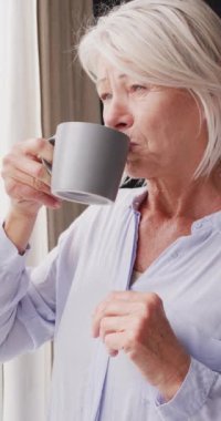 Yatak odasında pencereden bakan mutlu, yaşlı, beyaz bir kadının dikey videosu. Evde yalnız zaman geçirmek..