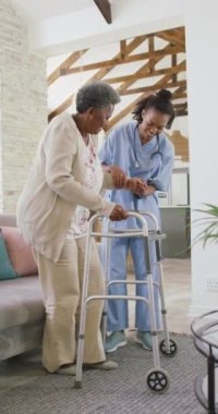 Yaşlı Afrikalı Amerikalı bir kadının kadın fizyoterapistle evde konuştuğu dikey bir video. Üst düzey sağlık ve tıbbi fizyoterapi tedavisi.