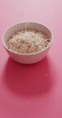 Tuzlu tuz kristallerinin pembe arka planda kaseye düşüşünün dikey videosu. yemek malzemeleri, yemek, lezzet ve besin.