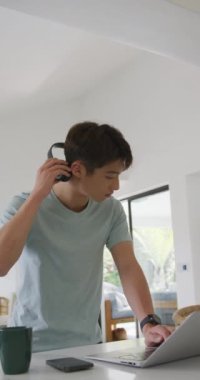 Asyalı bir gencin oturma odasında dizüstü bilgisayar kullandığının dikey videosu. Evde yalnız zaman geçirmek..