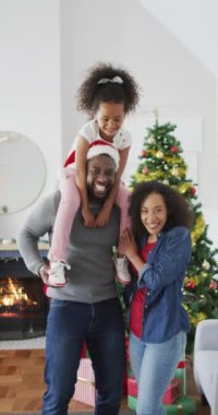 Afro-Amerikan bir ailenin kameraya bakarken dikey görüntüsü, babasının omuzlarında oturan kızı. Aile ve Noel konsepti.