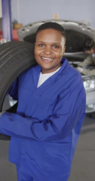 幸せなアフリカ系アメリカ人女性の車の整備士の笑顔と保持タイヤの垂直ビデオ 車修理工場で働き女性らしい小さなビジネスコンセプトを運営しています — ストック動画