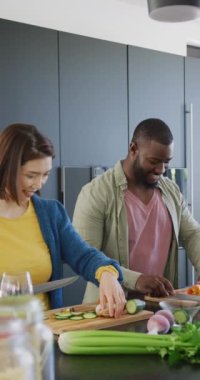 Vertical video of happy diverse couple preparing food together in kitchen. Domestic life, togetherness, health, happiness and inclusivity concept.