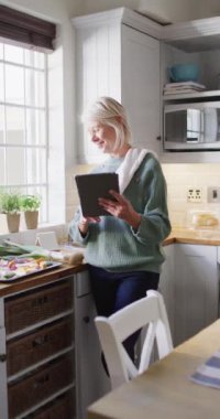 Mutfakta tablet kullanarak yemek hazırlayan mutlu, beyaz bir kadının dikey videosu. Evde yalnız zaman geçirmek..