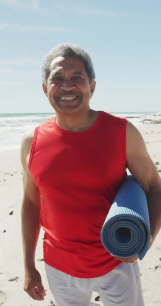 日当たりの良いビーチでヨガマットと幸せな出産の男の肖像画の垂直ビデオ 健康的で活発な退職ビーチの休日 — ストック動画