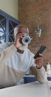 Afro-Amerikan bir adamın mutfakta kahve içerken akıllı telefonunu kullanmasının dikey görüntüsü. Yaşlılar ve teknoloji konsepti.