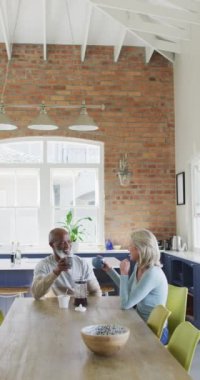 Son sınıf melez bir çiftin yemek masasında kahve içerken, gülerken ve gülümserken dikey görüntüsü. Yaşlılar ve ev hayatı kavramı.