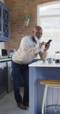 Afrika kökenli yaşlı bir adamın akıllı telefonuyla bir mutfak adasına eğildiği dikey bir fotoğraf. Yaşlılar ve teknoloji konsepti.