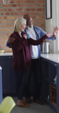 Pencereye el sallayan ve evde gülümseyen yaşlı bir çiftin dikey çekimi. Yaşlılar ve ev hayatı kavramı.