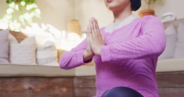 Evde yoga meditasyon nilüfer pozisyonunda oturan tesettürlü sakin melez kadının videosu. Mutluluk, sağlık, fitness, kapsayıcılık ve ev hayatı.